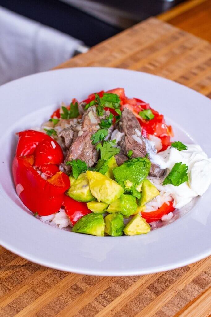 side view of oven baked steak fajitas in a white bowl topped with tomatoes, diced avocado, and sour cream