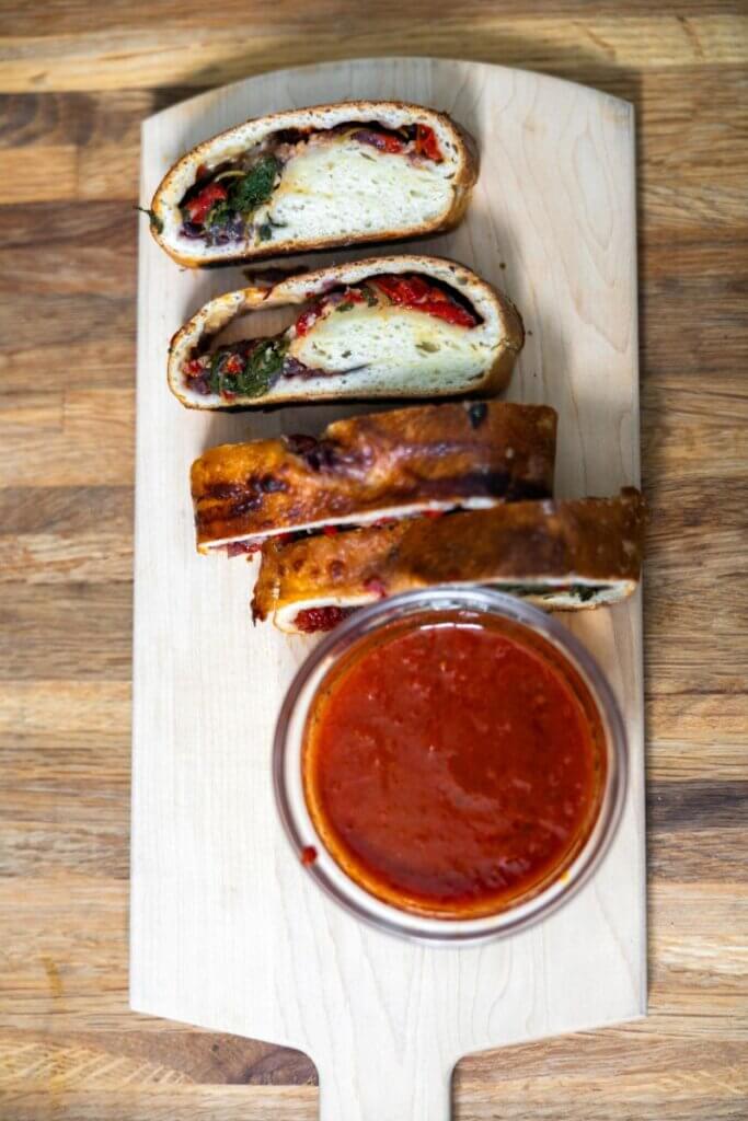 Homemade vegetable stromboli sliced next to marinara on a wooden cutting board.